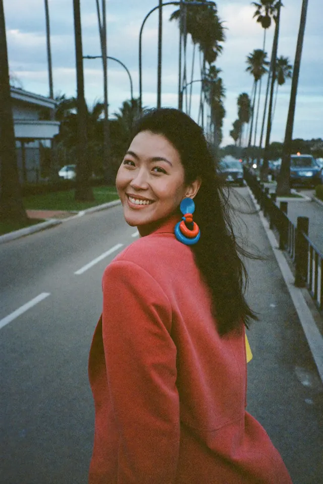 A woman in a coral blazer glances back over her shoulder on a palm-lined boulevard.