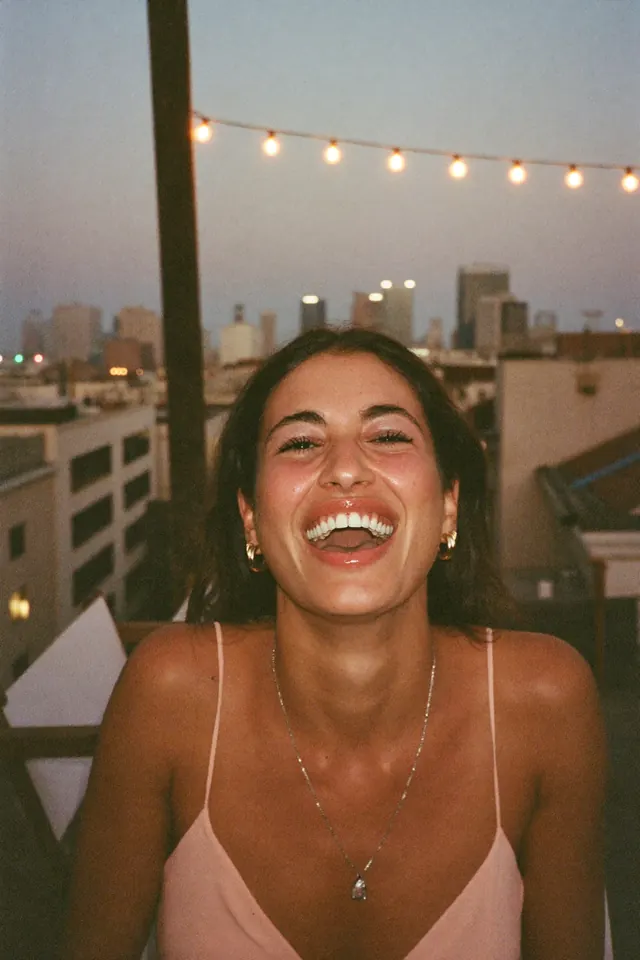 A woman laughs out loud on a rooftop terrace at dusk, string lights and the city behind her.